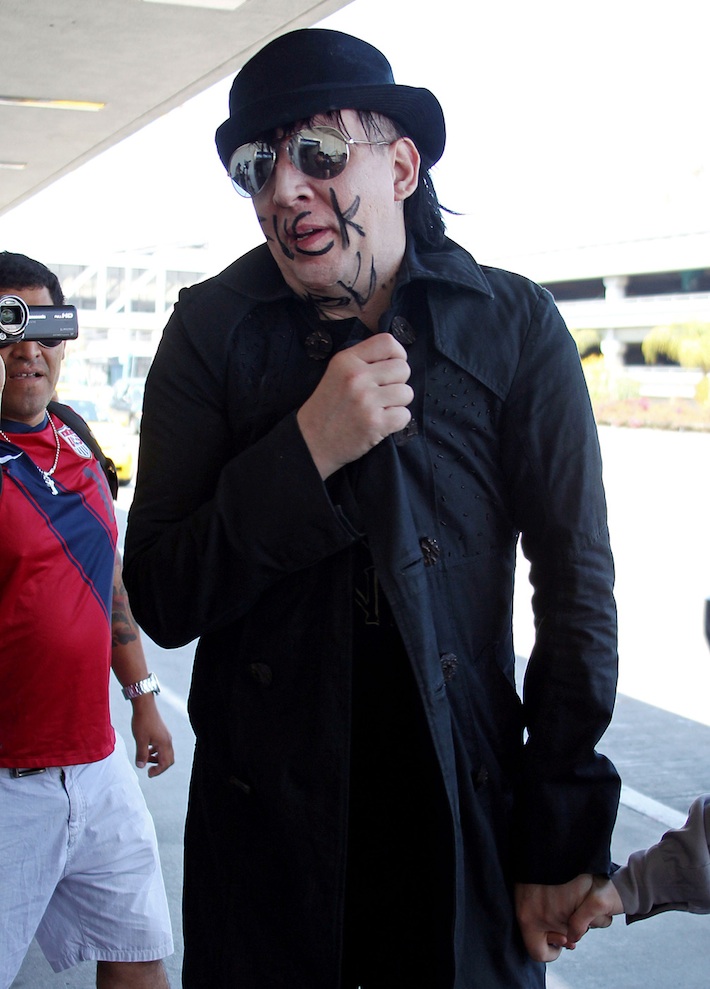 Marilyn Manson sunglasses and top hat writes f*ck you on his face and neck walking with his girlfriend